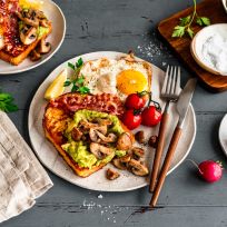 Ein Frühstücksteller mit Spiegeleiern, Speck, Herzhafter Frenchtoast mit Avocado und sautierten Champignons, Kirschtomaten und einer Zitronenscheibe. Daneben liegen Radieschen, Salz, eine Serviette und Besteck auf einem grauen Holztisch.