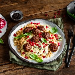 Ein Teller mit orientalisch gewürzten Fleischbällchen auf cremiger Frischkäsesauce und Couscous, garniert mit frischen Basilikumblättern und Granatapfelkernen, steht auf einem Holztisch. Daneben stehen Granatapfelhälften, eine kleine Schale mit Kernen und Besteck.