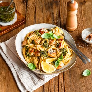 Ein Teller Pasta mit Hühnchen, frischem Pesto, Kirschtomaten und Basilikum, garniert mit Zitronenscheiben. Daneben stehen auf einem Holztisch ein Glas Pesto, eine Pfeffermühle, Zitronenspalten und ein Schälchen mit Salz.