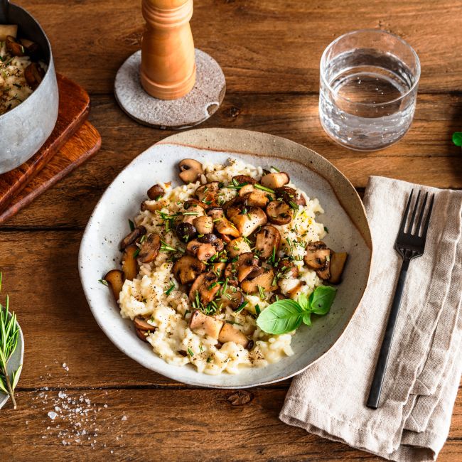 Auf einem Holztisch steht ein Teller mit cremigem Risotto, garniert mit sautierten Pilzen und Kräutern, neben einer Gabel und einer Serviette, einer Schüssel mit Salz, einem Glas Wasser, einem Topf mit Pilzen und einem kleinen Teller mit frischem Gemüse.