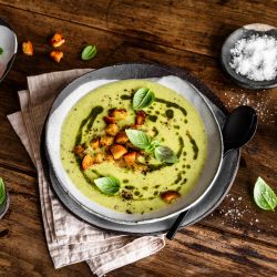 Eine Schüssel cremige Zucchinisuppe, garniert mit Croutons, Basilikumblättern und einem Spritzer Kräuteröl, steht auf einem Holztisch. Daneben liegen weitere Croutons, Salz und Basilikum. Ein Löffel liegt auf dem Rand der Schüssel.