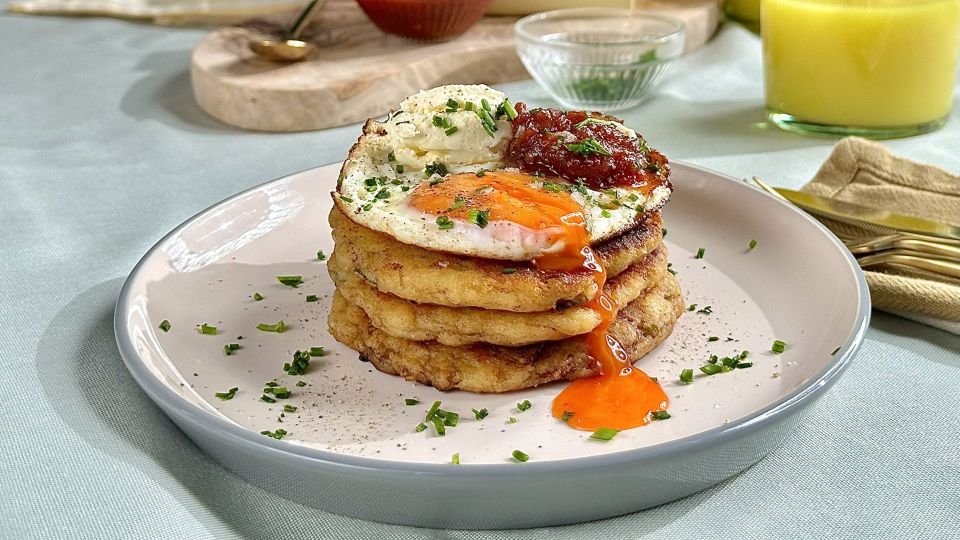 Ein Stapel herzhafter Kartoffelpüreepuffer mit einem Spiegelei, einem Klecks Sahne, roter Sauce und gehackten Kräutern auf einem weißen Teller. Das zerbrochene, flüssige Eigelb verleiht dem Ganzen eine besondere Note. Im Hintergrund steht ein Glas Orangensaft.