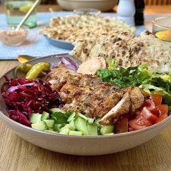 Eine Hähnchenbowl gefüllt mit gegrilltem Hähnchen, Gurken, Tomaten, Salat, Rotkohl, eingelegter Paprika, roten Zwiebeln und Sauce wird mit Naan-Brot serviert. Ein erfrischendes Zitronengetränk und weitere Gerichte stehen im Hintergrund.