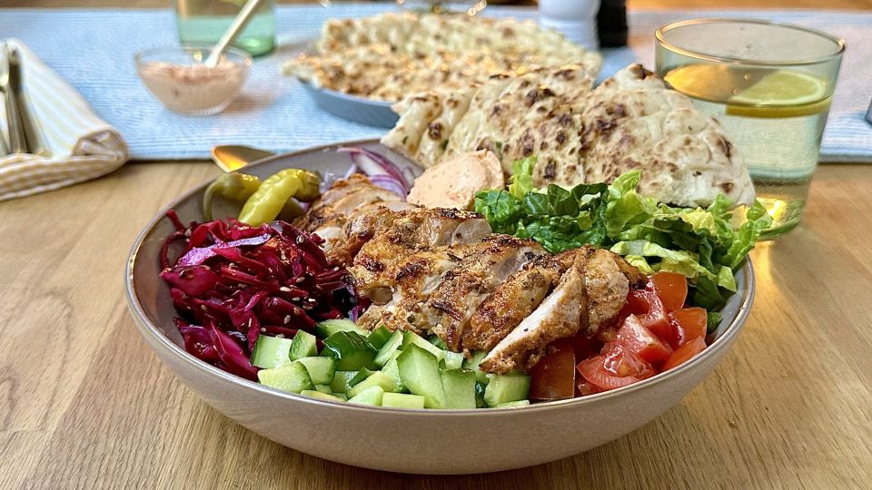 Eine Hähnchenbowl gefüllt mit gegrilltem Hähnchen, Gurken, Tomaten, Salat, Rotkohl, eingelegter Paprika, roten Zwiebeln und Sauce wird mit Naan-Brot serviert. Ein erfrischendes Zitronengetränk und weitere Gerichte stehen im Hintergrund.