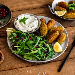 Ein Teller mit knusprigen Falafeln, Zitronenspalten und frischem grünen Salat mit Granatapfelkernen wird zusammen mit einer Schüssel cremigem Dip serviert. Eine Gabel, Petersilie, Pfeffer und zusätzliche Falafel werden auf einem Holztisch angerichtet.