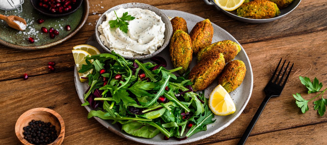 Ein Teller mit knusprigen Falafeln, Zitronenspalten und frischem grünen Salat mit Granatapfelkernen wird zusammen mit einer Schüssel cremigem Dip serviert. Eine Gabel, Petersilie, Pfeffer und zusätzliche Falafel werden auf einem Holztisch angerichtet.