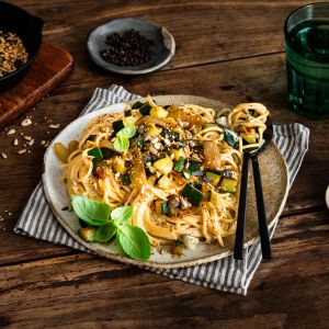 Ein Teller Pasta mit geröstete Zucchini, frischem Basilikum und Samen wird auf einer gestreiften Serviette mit Besteck serviert. Daneben stehen ein Glas Wasser, eine Schale mit Salz, in einer Pfanne geröstete Samen und ein kleiner Teller mit Basilikumblättern.