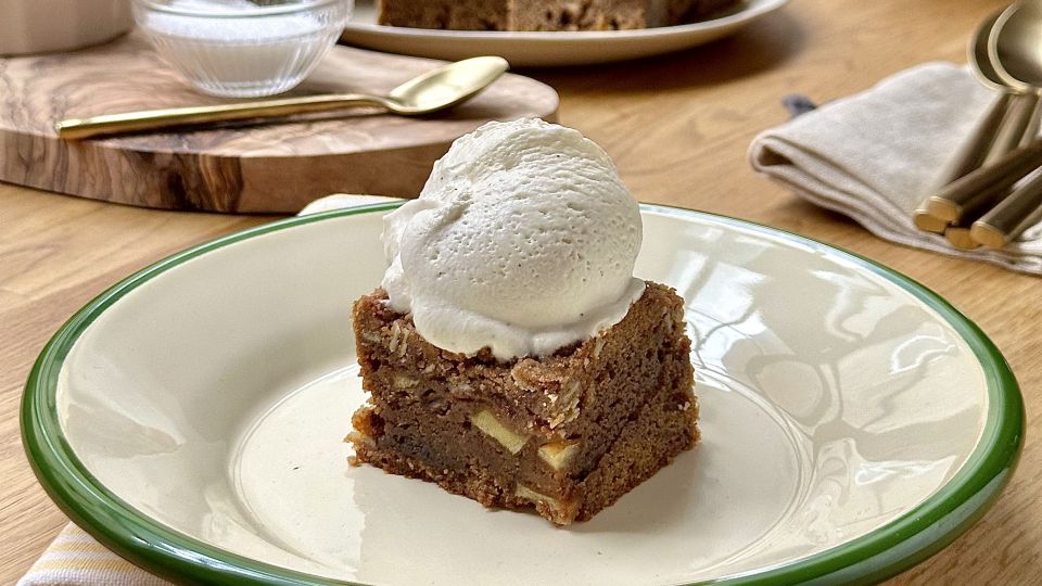 Ein quadratisches Stück Apfelblondies-Kuchen mit sichtbaren Apfelstücken wird auf einem Teller serviert, garniert mit Vanilleeis. Im Hintergrund stehen eine Schüssel mit Zucker und Besteck auf einem Holztisch.