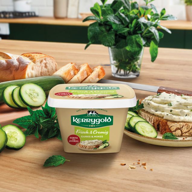 Ein Behälter mit Kerrygold Frischkäse steht auf einem Holztisch, mit Gurkenscheiben, einem Laib Brot, frischen Kräutern und einer mit Frischkäse belegten Brotscheibe. Im Hintergrund steht frischer Basilikum.