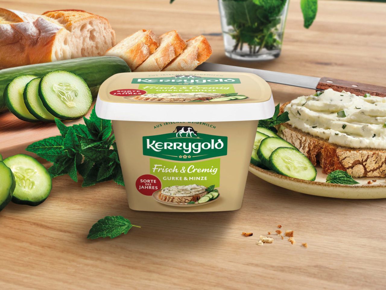 Ein Behälter mit Kerrygold Frischkäse steht auf einem Holztisch, mit Gurkenscheiben, einem Laib Brot, frischen Kräutern und einer mit Frischkäse belegten Brotscheibe. Im Hintergrund steht frischer Basilikum.