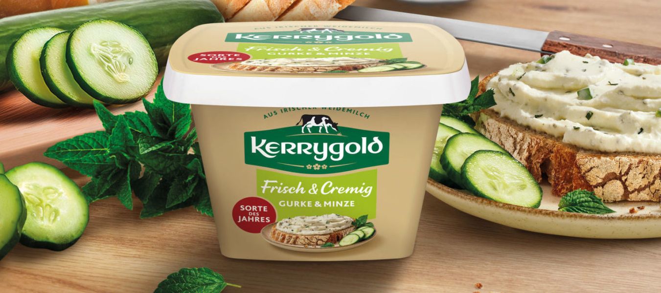 Ein Behälter mit Kerrygold Frischkäse steht auf einem Holztisch, mit Gurkenscheiben, einem Laib Brot, frischen Kräutern und einer mit Frischkäse belegten Brotscheibe. Im Hintergrund steht frischer Basilikum.