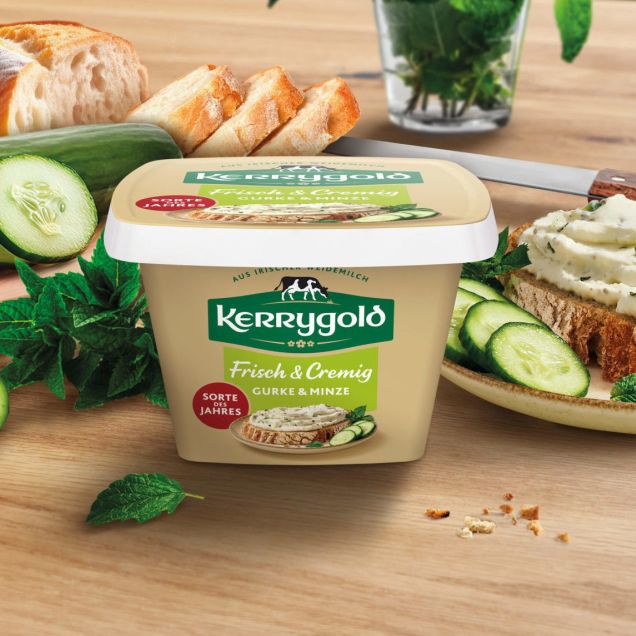 Ein Behälter mit Kerrygold Frischkäse steht auf einem Holztisch, mit Gurkenscheiben, einem Laib Brot, frischen Kräutern und einer mit Frischkäse belegten Brotscheibe. Im Hintergrund steht frischer Basilikum.