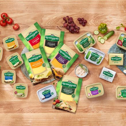 Eine Produktübersicht der Kerrygold Käse- und Butterprodukte ist auf einem Holztisch mit Brot, Trauben, geschnittenem Gemüse, geriebenem Käse und Kräutern arrangiert. Die Verpackung ist in klassischen Grün- und Gelbtönen gehalten.