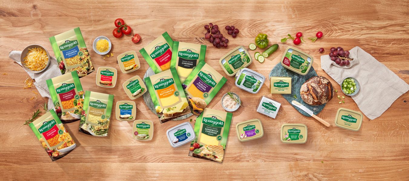 Eine Produktübersicht der Kerrygold Käse- und Butterprodukte ist auf einem Holztisch mit Brot, Trauben, geschnittenem Gemüse, geriebenem Käse und Kräutern arrangiert. Die Verpackung ist in klassischen Grün- und Gelbtönen gehalten.