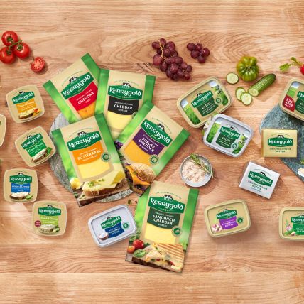Eine Produktübersicht der Kerrygold Käse- und Butterprodukte ist auf einem Holztisch mit Brot, Trauben, geschnittenem Gemüse, geriebenem Käse und Kräutern arrangiert. Die Verpackung ist in klassischen Grün- und Gelbtönen gehalten.