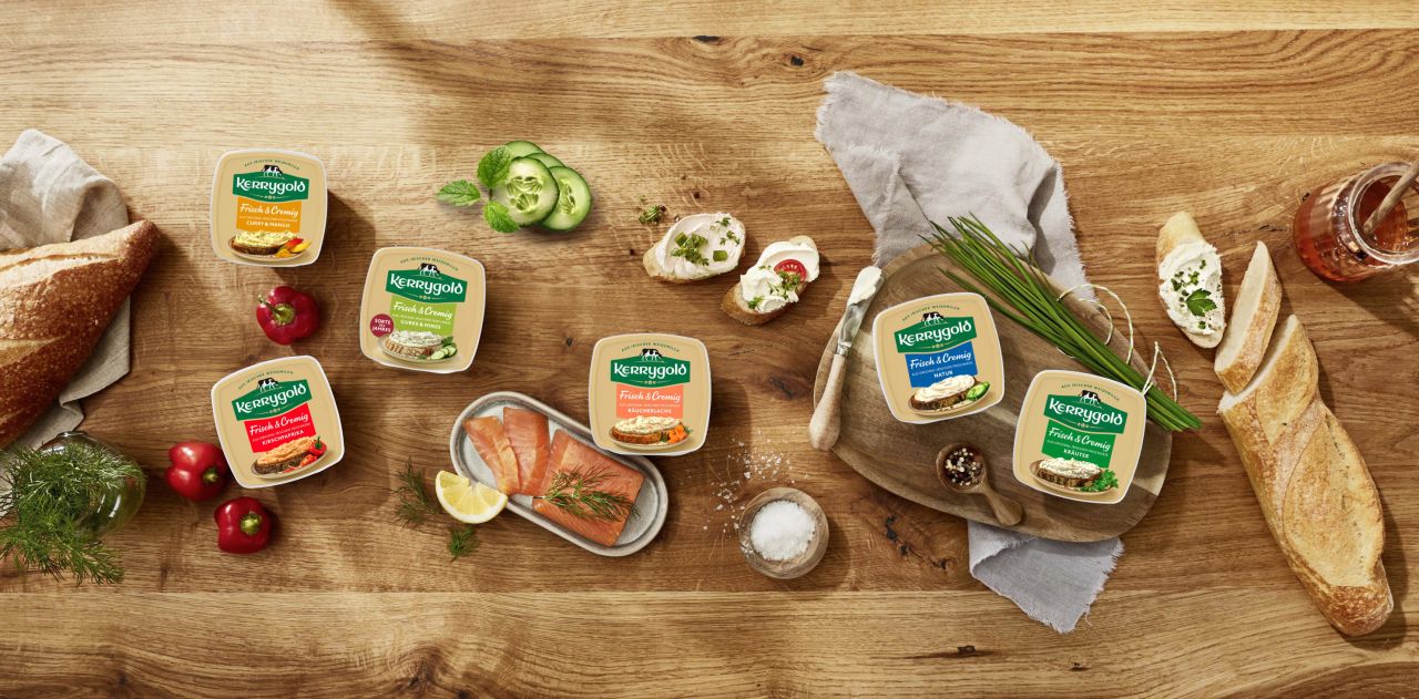 Ein Holztisch mit einer Produktübersicht verschiedener Frischkäsesorten im Becher, geschnittenem Brot, Baguette, Tomaten, Kräutern, Gurken, Räucherlachs und einem Glas Honig, frisch und appetitlich angerichtet.