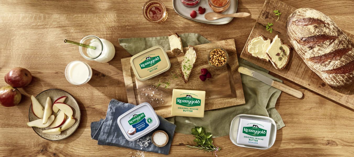 Ein Holztisch mit verschiedenen Kerrygold Butter-Produkten, Brot, Apfelscheiben, Nüssen, Marmeladengläsern, einem Glas Milch und einem Buttermesser sorgt für eine rustikale Frühstücks- oder Snackszene.