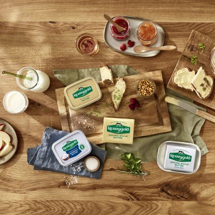 Ein Holztisch mit verschiedenen Kerrygold Butter-Produkten, Brot, Apfelscheiben, Nüssen, Marmeladengläsern, einem Glas Milch und einem Buttermesser sorgt für eine rustikale Frühstücks- oder Snackszene.