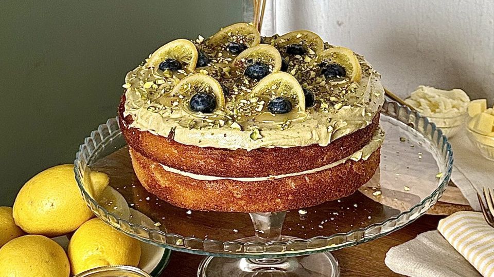 Ein zweischichtiger Zitronen-Pistazien-Kuchen mit cremigem Zuckerguss, garniert mit Zitronenscheiben, Blaubeeren und gehackten Pistazien, präsentiert auf einem Glaskuchenständer neben ganzen Zitronen und einem Teller mit Gabeln.
