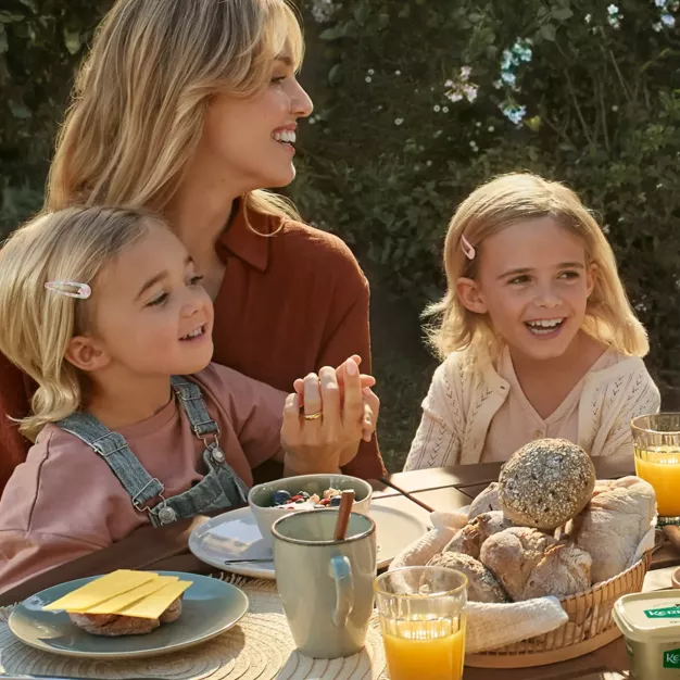 Eine Frau und zwei kleine Kinder sitzen lächelnd und händchenhaltend an einem Tisch im Freien. Der Tisch ist mit Brot, Käse, Müsli, Obst und Gläsern Orangensaft gedeckt. Sonnenlicht und Grün umgeben sie.
