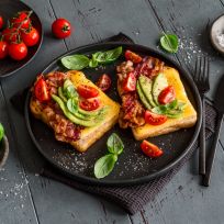 Auf einem schwarzen Teller liegen zwei Stullen-Generator-Toasts mit Speck, Avocadoscheiben, Kirschtomaten und frischem Basilikum, umgeben von Basilikumblättern, Kirschtomaten an der Rebe, einer Schale mit Salz und Besteck auf einem blauen Holztisch.