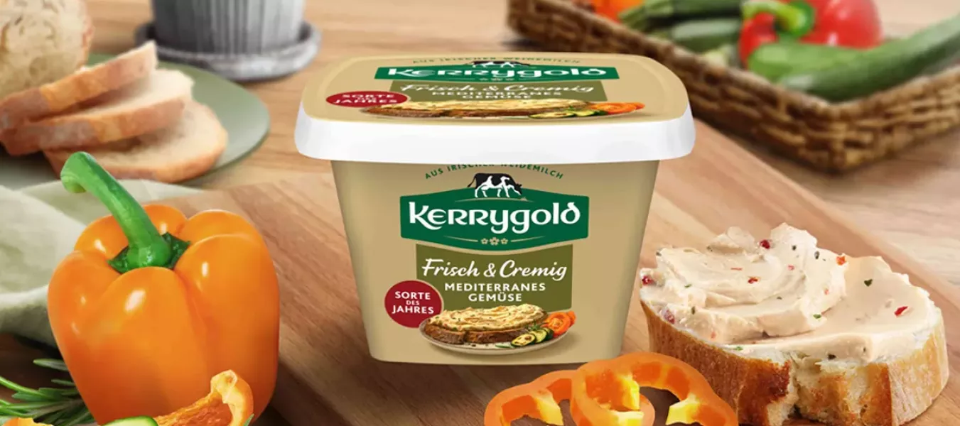 Ein Behälter mit Kerrygold Mediterranes Gemüse, der Sorte des Jahres, steht auf einem hölzernen Schneidebrett, umgeben von geschnittenem Brot, einem Messer, gelben Paprikascheiben, Zucchini und einem Korb mit verschiedenem Gemüse im Hintergrund.