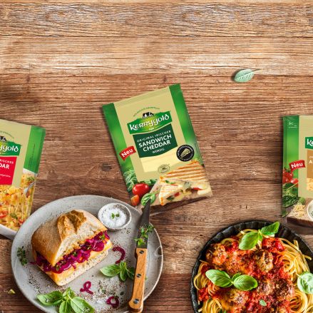 Vier Sorten Kerrygold-Käse, darunter geschnittener Cheddar und geriebener Käse, werden auf einem Holztisch mit zubereiteten Gerichten wie Pasta, Loaded Fries, einem Sandwich und gefülltem Brot für den ultimativen Käsegenuss präsentiert.