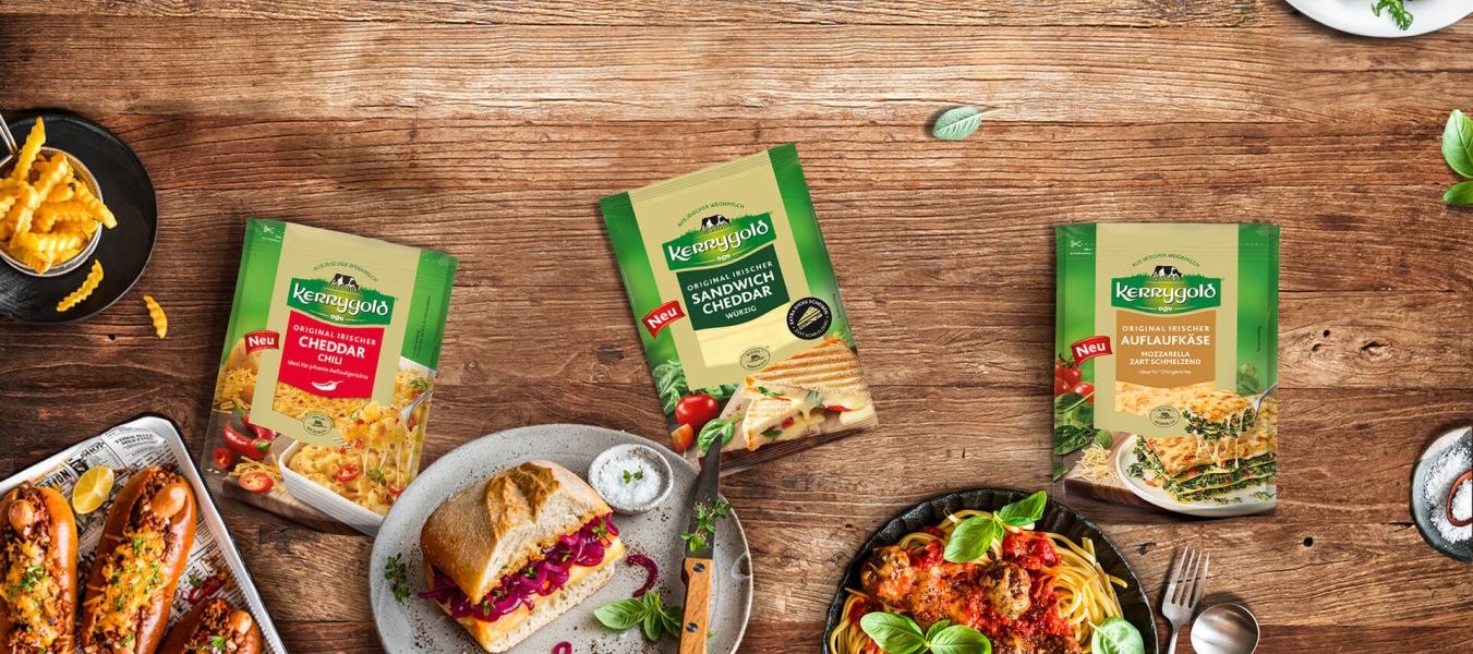Vier Sorten Kerrygold-Käse, darunter geschnittener Cheddar und geriebener Käse, werden auf einem Holztisch mit zubereiteten Gerichten wie Pasta, Loaded Fries, einem Sandwich und gefülltem Brot für den ultimativen Käsegenuss präsentiert.