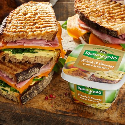Gegrillte Sandwiches mit Schinken, Käse und Gemüse sind auf einem Holzbrett neben einem Behälter mit Kerrygold Frischkäse Curry & Mango, der Sorte des Jahres – für exotischen Geschmack, gestapelt. Im Hintergrund sind Brot und Spinat zu sehen.
