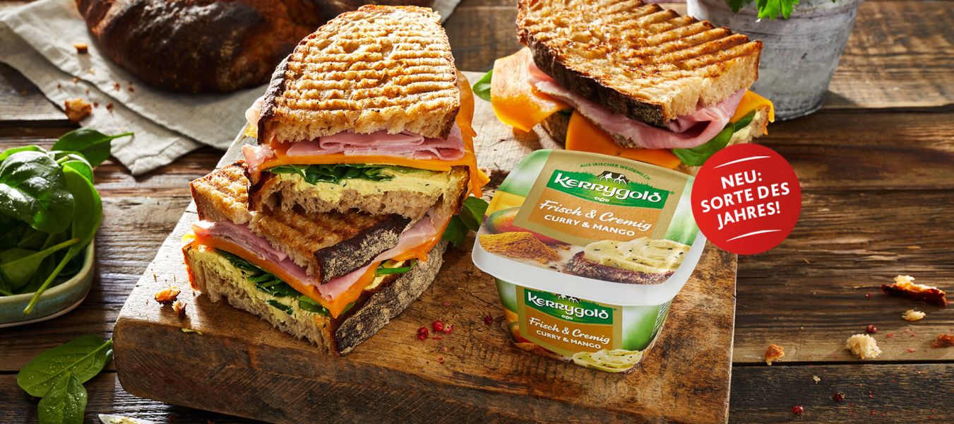 Gegrillte Sandwiches mit Schinken, Käse und Gemüse sind auf einem Holzbrett neben einem Behälter mit Kerrygold Frischkäse Curry & Mango, der Sorte des Jahres – für exotischen Geschmack, gestapelt. Im Hintergrund sind Brot und Spinat zu sehen.