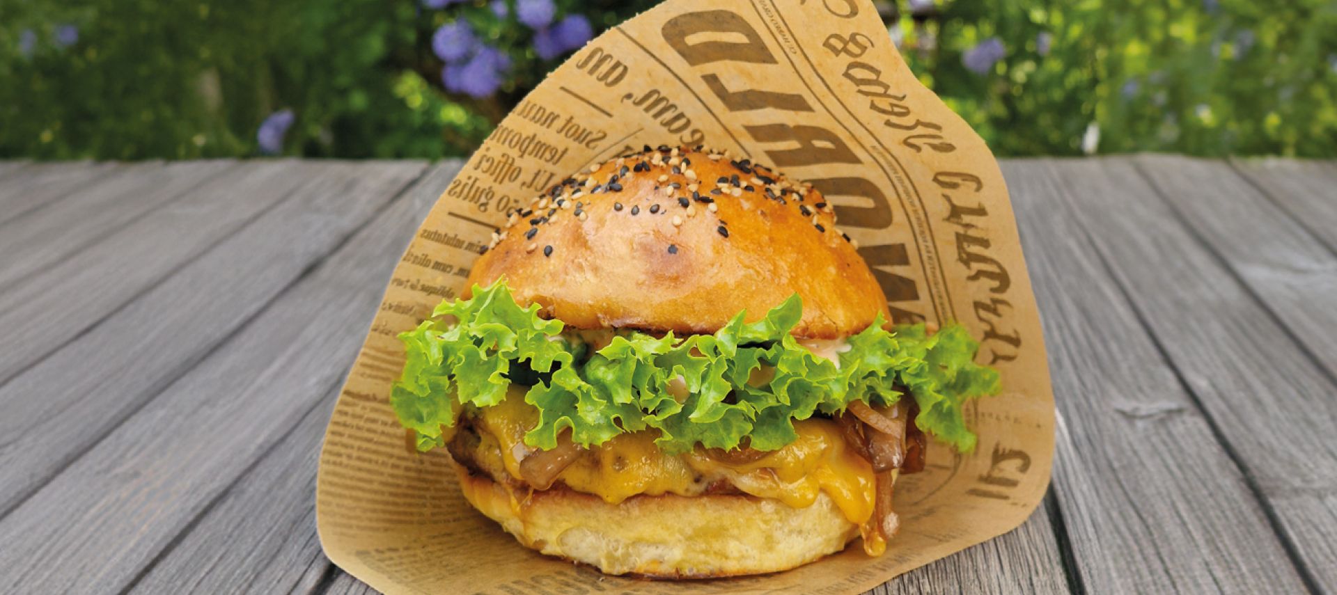 Ein leckerer Burger mit Salat und einem Sesambrötchen, eingewickelt in bedrucktes Papier, liegt auf einer Holzfläche im Freien, umgeben von grünem Laub und violetten Blüten im leicht verschwommenen Hintergrund.