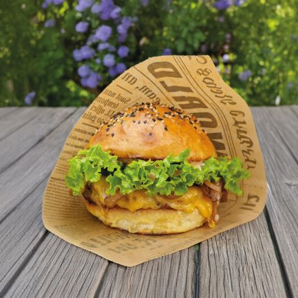Ein leckerer Burger mit Salat und einem Sesambrötchen, eingewickelt in bedrucktes Papier, liegt auf einer Holzfläche im Freien, umgeben von grünem Laub und violetten Blüten im leicht verschwommenen Hintergrund.