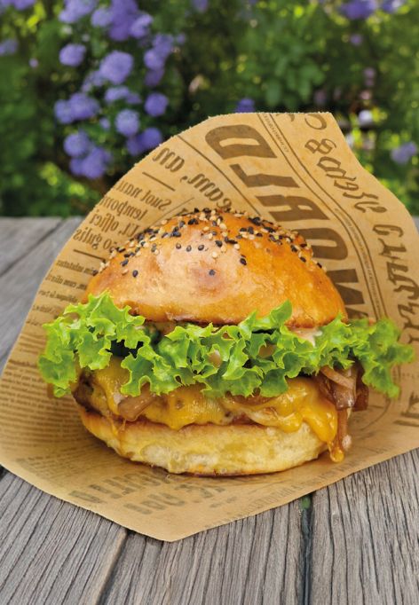 Ein leckerer Burger mit Salat und einem Sesambrötchen, eingewickelt in bedrucktes Papier, liegt auf einer Holzfläche im Freien, umgeben von grünem Laub und violetten Blüten im leicht verschwommenen Hintergrund.