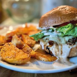 Der Honey Explosion Burger mit Salat, Tomaten und cremiger Sauce wird auf einem weißen Teller neben goldbraunen, gewürzten Kartoffelecken serviert. Der sanft verschwommene Hintergrund unterstreicht dieses schmackhafte Gericht.