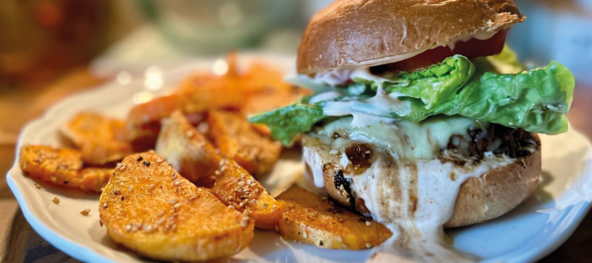 Der Honey Explosion Burger mit Salat, Tomaten und cremiger Sauce wird auf einem weißen Teller neben goldbraunen, gewürzten Kartoffelecken serviert. Der sanft verschwommene Hintergrund unterstreicht dieses schmackhafte Gericht.