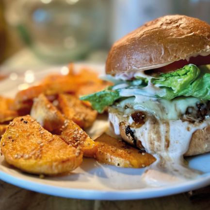 Der Honey Explosion Burger mit Salat, Tomaten und cremiger Sauce wird auf einem weißen Teller neben goldbraunen, gewürzten Kartoffelecken serviert. Der sanft verschwommene Hintergrund unterstreicht dieses schmackhafte Gericht.