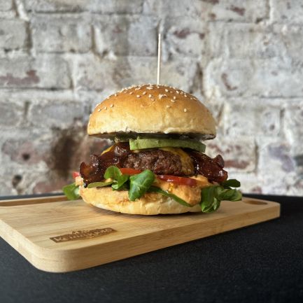 Der Männi Burger ist ein Gourmet-Burger mit Salat, Tomaten, Speck, Avocado und Sauce in einem Sesambrötchen, serviert auf einem Holzbrett vor einem rustikalen Backsteinmauerhintergrund.