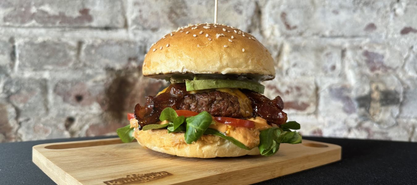 Der Männi Burger ist ein Gourmet-Burger mit Salat, Tomaten, Speck, Avocado und Sauce in einem Sesambrötchen, serviert auf einem Holzbrett vor einem rustikalen Backsteinmauerhintergrund.