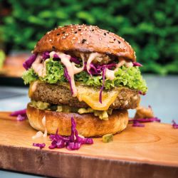 Eine Nahaufnahme eines pflanzlichen Burgers der nächsten Generation mit Salat, zerkleinertem Rotkohl, Käse, Gurken und Soße auf einem Sesambrötchen, serviert auf einem Holzbrett mit Grünpflanzen im unscharfen Hintergrund.