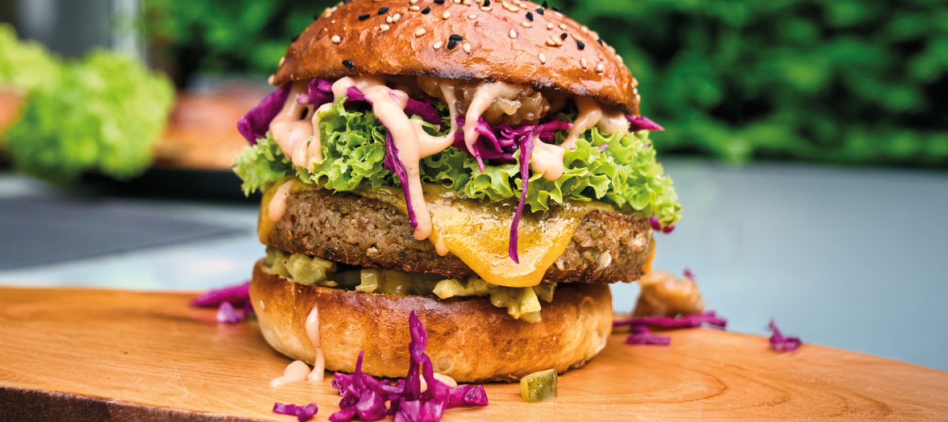 Eine Nahaufnahme eines pflanzlichen Burgers der nächsten Generation mit Salat, zerkleinertem Rotkohl, Käse, Gurken und Soße auf einem Sesambrötchen, serviert auf einem Holzbrett mit Grünpflanzen im unscharfen Hintergrund.