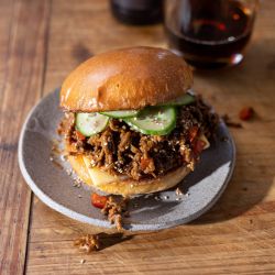Ein Sloppy Joe Burger mit Gurkenscheiben liegt auf einem grauen Teller auf einem Holztisch. Im Hintergrund sind ein Getränk und eine Flasche unscharf zu sehen. Auf dem Teller sind etwas zerkleinertes Rindfleisch und Sesamsamen verstreut.