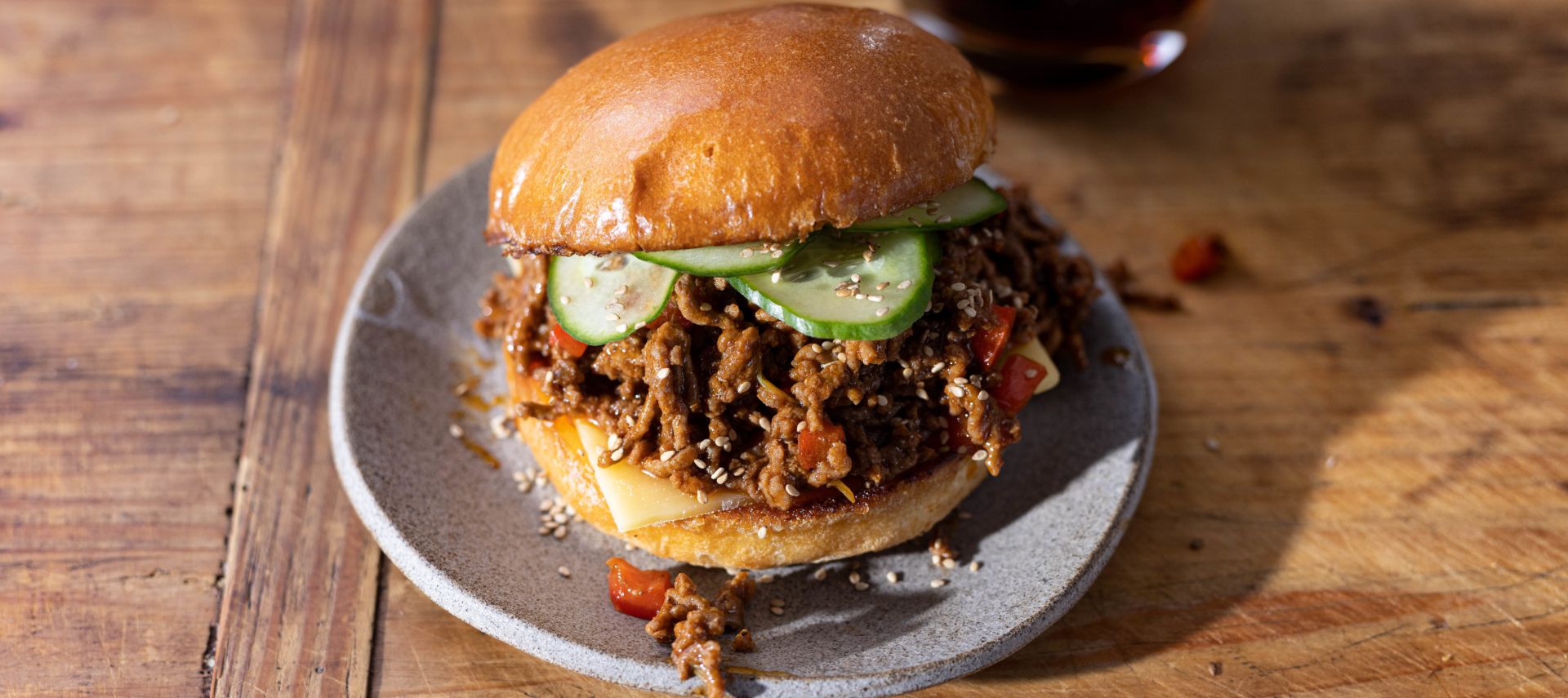 Ein Sloppy Joe Burger mit Gurkenscheiben liegt auf einem grauen Teller auf einem Holztisch. Im Hintergrund sind ein Getränk und eine Flasche unscharf zu sehen. Auf dem Teller sind etwas zerkleinertes Rindfleisch und Sesamsamen verstreut.