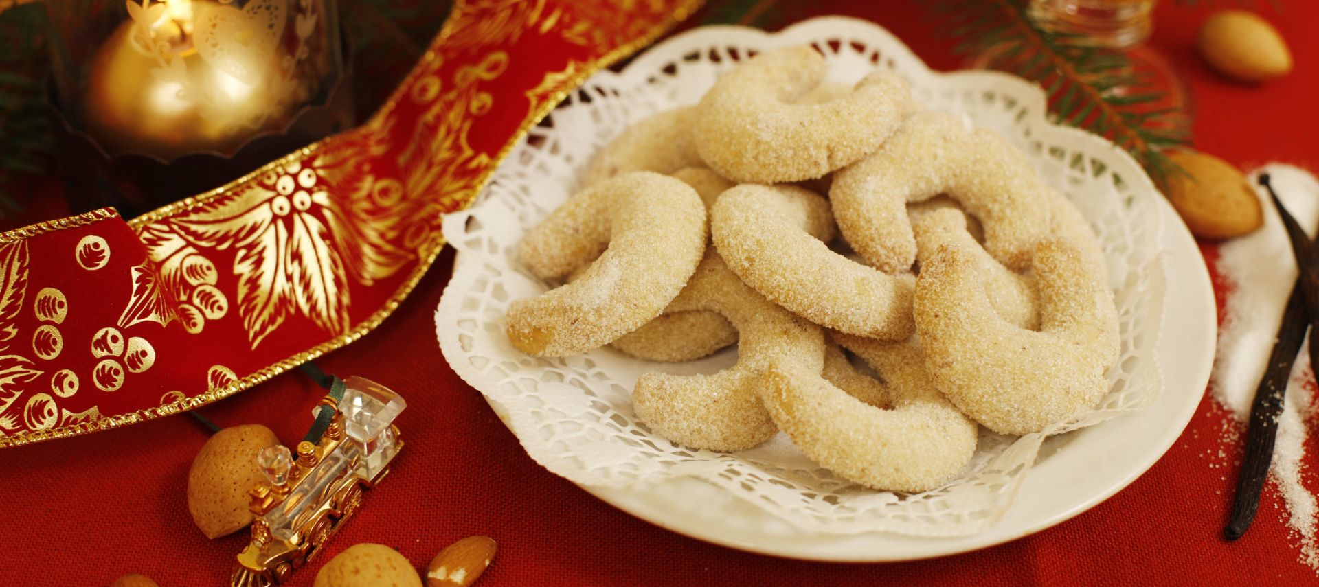 Ein Teller mit Vanillekipferl, traditionellen österreichischen Vanillekipferln, die mit Puderzucker bestäubt sind, steht auf einer Spitzendeckchen inmitten von Mandeln, einem festlichen Tischtuch, einer brennenden Kerze und Weihnachtsdekorationen auf einer roten Tischdecke.