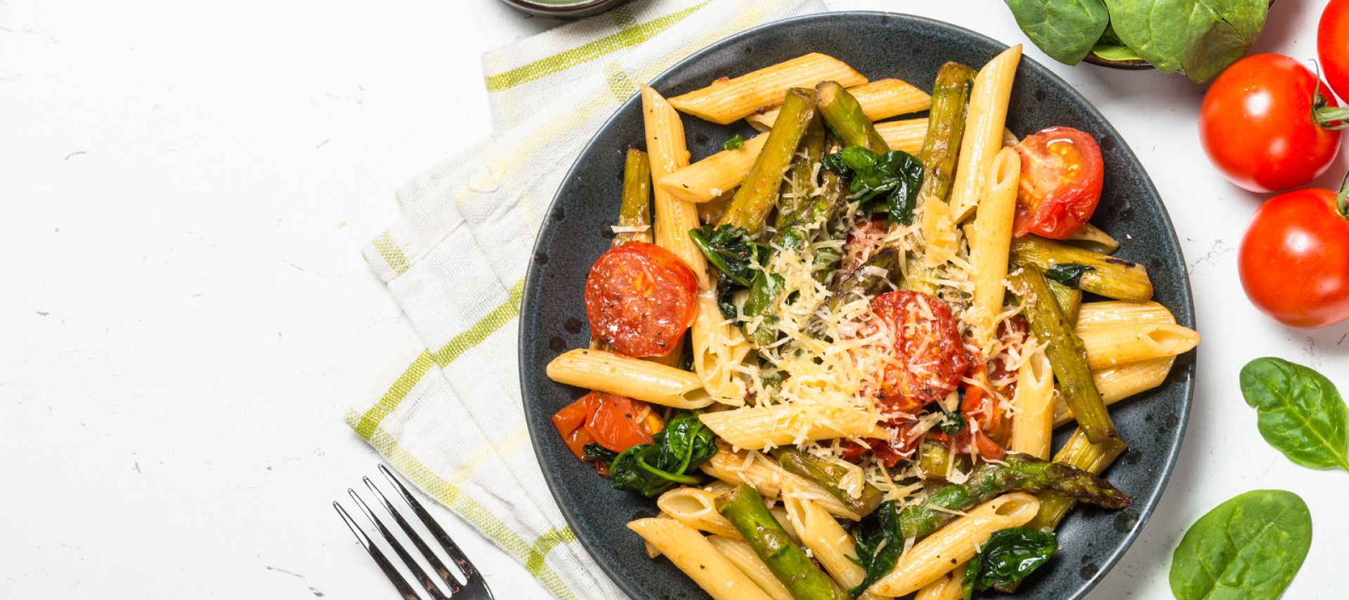Ein schwarzer Teller mit Penne-Nudeln, Kirschtomaten, Spargel, Spinat und geriebenem Käse steht auf einer Stoffserviette. Daneben liegen eine Gabel, eine Schüssel Käse, frische Spinatblätter und getrocknete Tomaten, verstreut auf einer weißen Fläche.