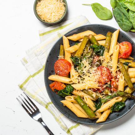 Ein schwarzer Teller mit Penne-Nudeln, Kirschtomaten, Spargel, Spinat und geriebenem Käse steht auf einer Stoffserviette. Daneben liegen eine Gabel, eine Schüssel Käse, frische Spinatblätter und getrocknete Tomaten, verstreut auf einer weißen Fläche.