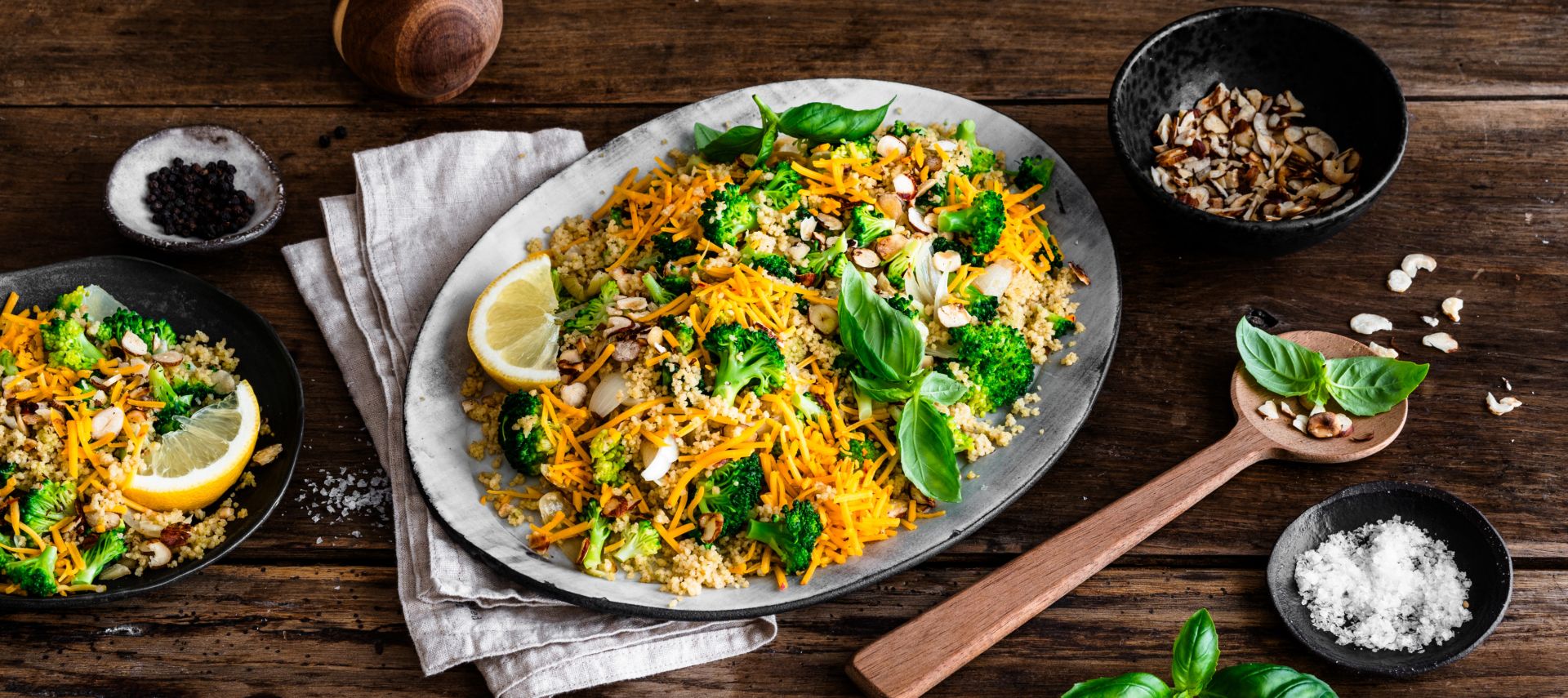 Eine Platte mit farbenfrohem Gemüsereis, inspiriert von Brokkoli-Couscous-Salat und garniert mit Nüssen, frischem Basilikum und Zitronenscheiben, steht auf einem rustikalen Holztisch neben Mandelblättchen, geriebenem Chili-Cheddar, einer Pfeffermühle und einem Holzlöffel.