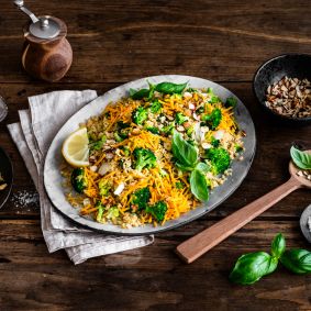 Eine Platte mit farbenfrohem Gemüsereis, inspiriert von Brokkoli-Couscous-Salat und garniert mit Nüssen, frischem Basilikum und Zitronenscheiben, steht auf einem rustikalen Holztisch neben Mandelblättchen, geriebenem Chili-Cheddar, einer Pfeffermühle und einem Holzlöffel.