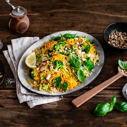 Eine Platte mit farbenfrohem Gemüsereis, inspiriert von Brokkoli-Couscous-Salat und garniert mit Nüssen, frischem Basilikum und Zitronenscheiben, steht auf einem rustikalen Holztisch neben Mandelblättchen, geriebenem Chili-Cheddar, einer Pfeffermühle und einem Holzlöffel.