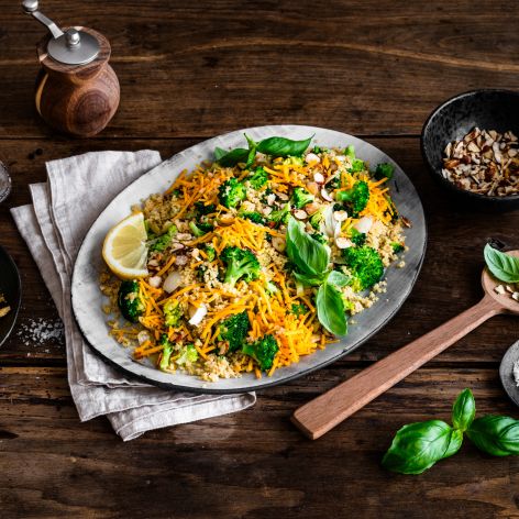 Eine Platte mit farbenfrohem Gemüsereis, inspiriert von Brokkoli-Couscous-Salat und garniert mit Nüssen, frischem Basilikum und Zitronenscheiben, steht auf einem rustikalen Holztisch neben Mandelblättchen, geriebenem Chili-Cheddar, einer Pfeffermühle und einem Holzlöffel.