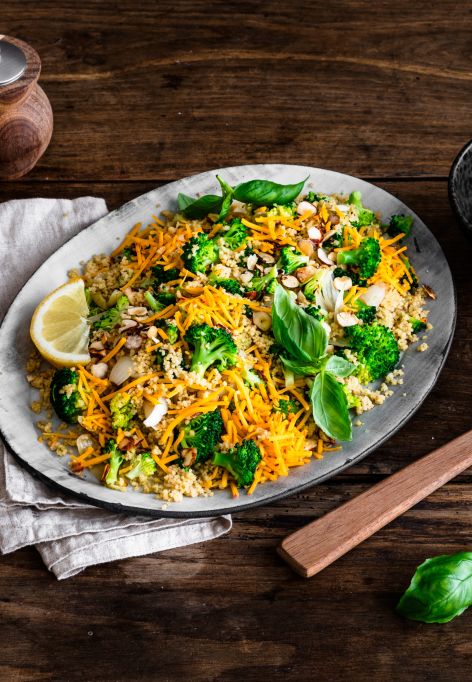 Eine Platte mit farbenfrohem Gemüsereis, inspiriert von Brokkoli-Couscous-Salat und garniert mit Nüssen, frischem Basilikum und Zitronenscheiben, steht auf einem rustikalen Holztisch neben Mandelblättchen, geriebenem Chili-Cheddar, einer Pfeffermühle und einem Holzlöffel.